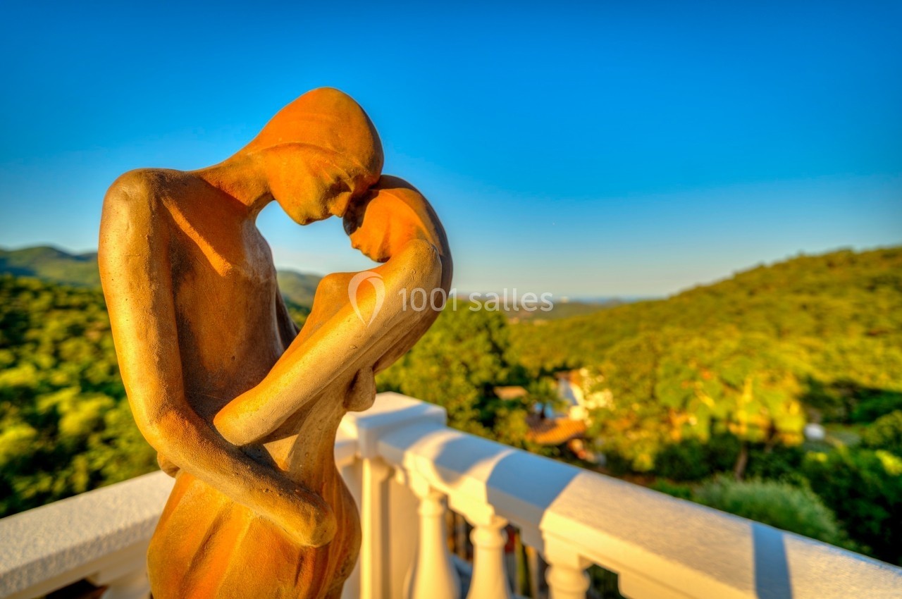 Statue en bronze représentant un couple s'enlaçant, placée sur une terrasse avec vue sur un paysage verdoyant.