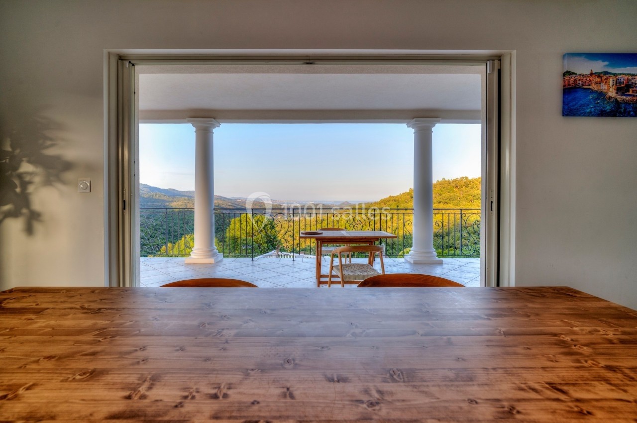Vue depuis une table en bois sur une terrasse avec balustrade, colonnes blanches et paysage vallonné en arrière-plan.