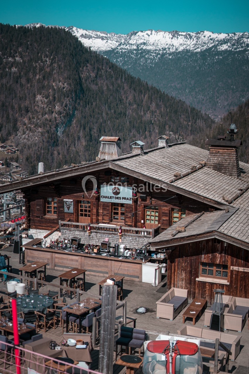 Chalet en bois avec terrasse aménagée, entouré de montagnes et de forêts sous un ciel dégagé.