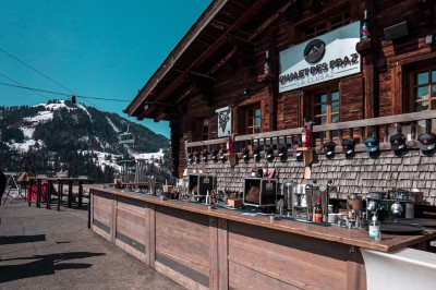 Une femme souriante se tient derrière une table en bois garnie de plats dans un chalet en bois éclairé chaleureusement.