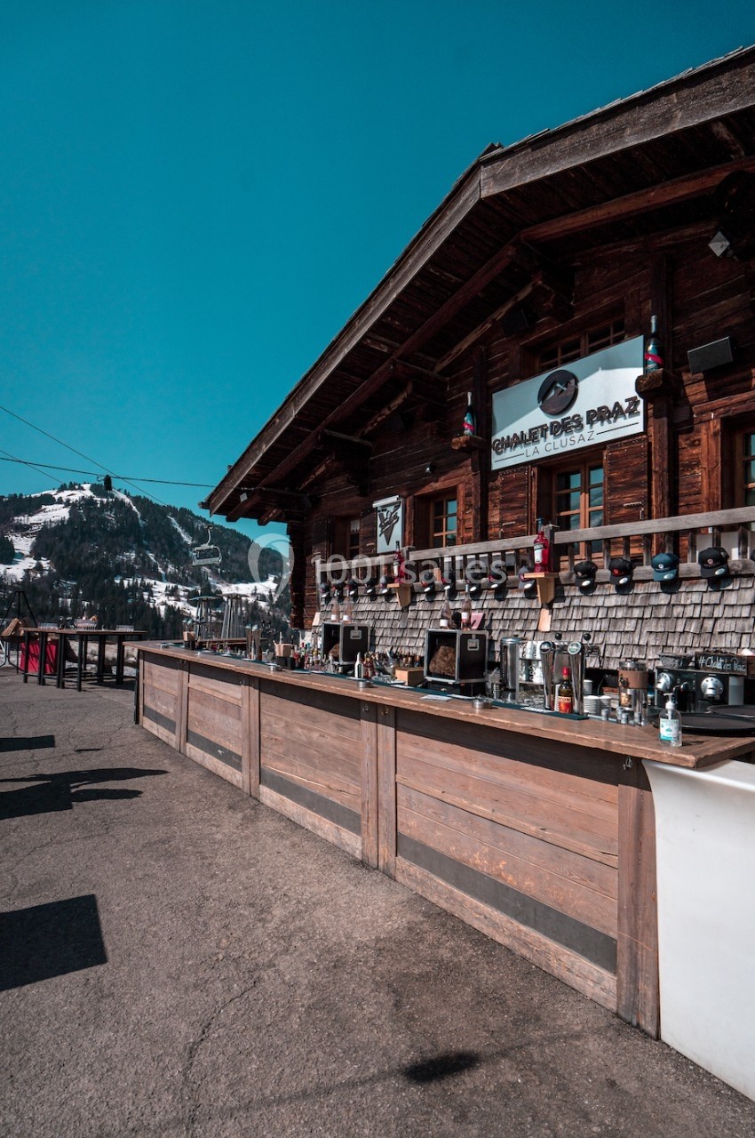 Chalet en bois avec terrasse extérieure, bar aménagé et vue sur des montagnes enneigées sous un ciel bleu.