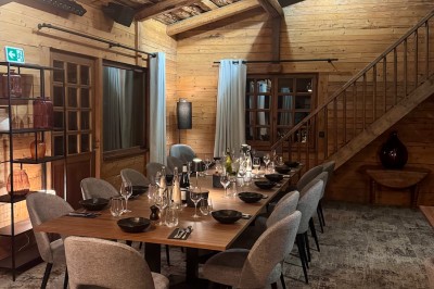 Une femme souriante se tient derrière une table en bois garnie de plats dans un chalet en bois éclairé chaleureusement.