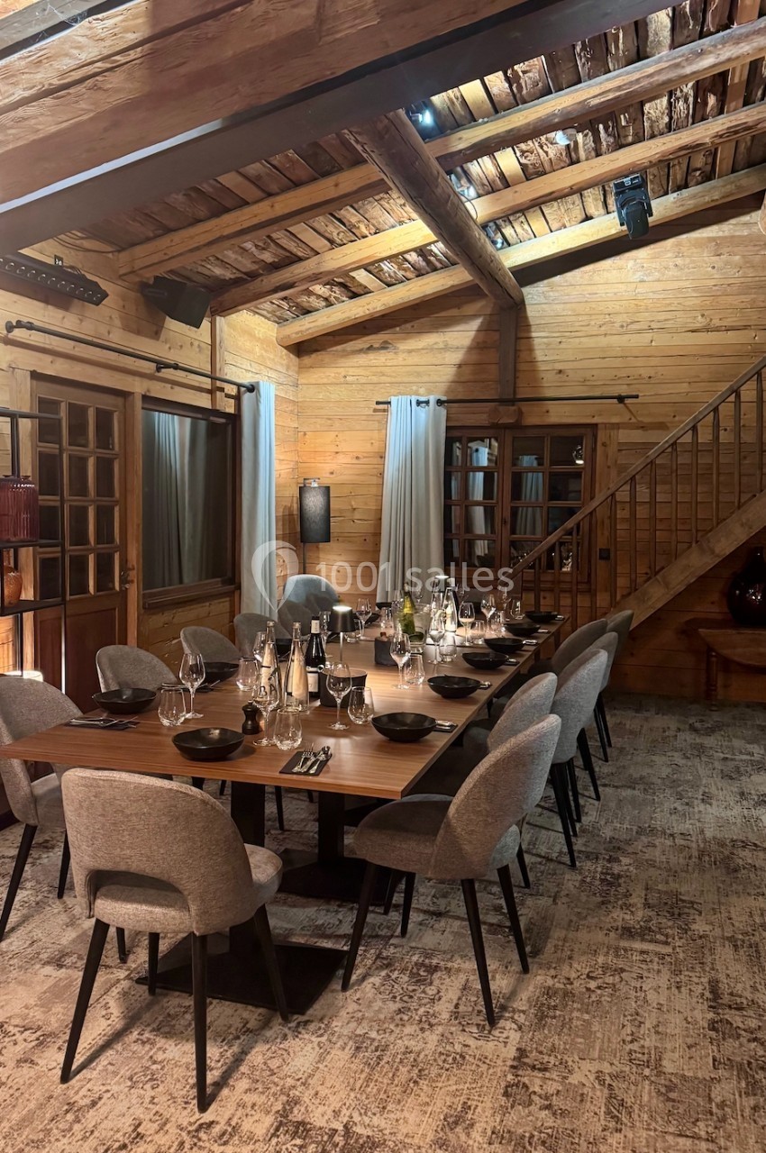 Salle à manger chaleureuse avec table en bois dressée, chaises grises, murs en bois et plafond avec poutres apparentes.