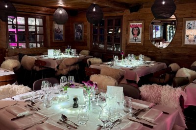 Une femme souriante se tient derrière une table en bois garnie de plats dans un chalet en bois éclairé chaleureusement.