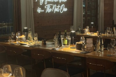 Une femme souriante se tient derrière une table en bois garnie de plats dans un chalet en bois éclairé chaleureusement.