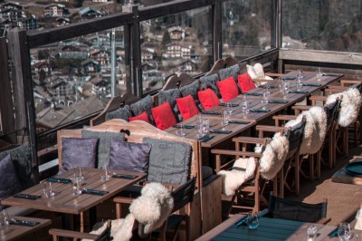 Une femme souriante se tient derrière une table en bois garnie de plats dans un chalet en bois éclairé chaleureusement.