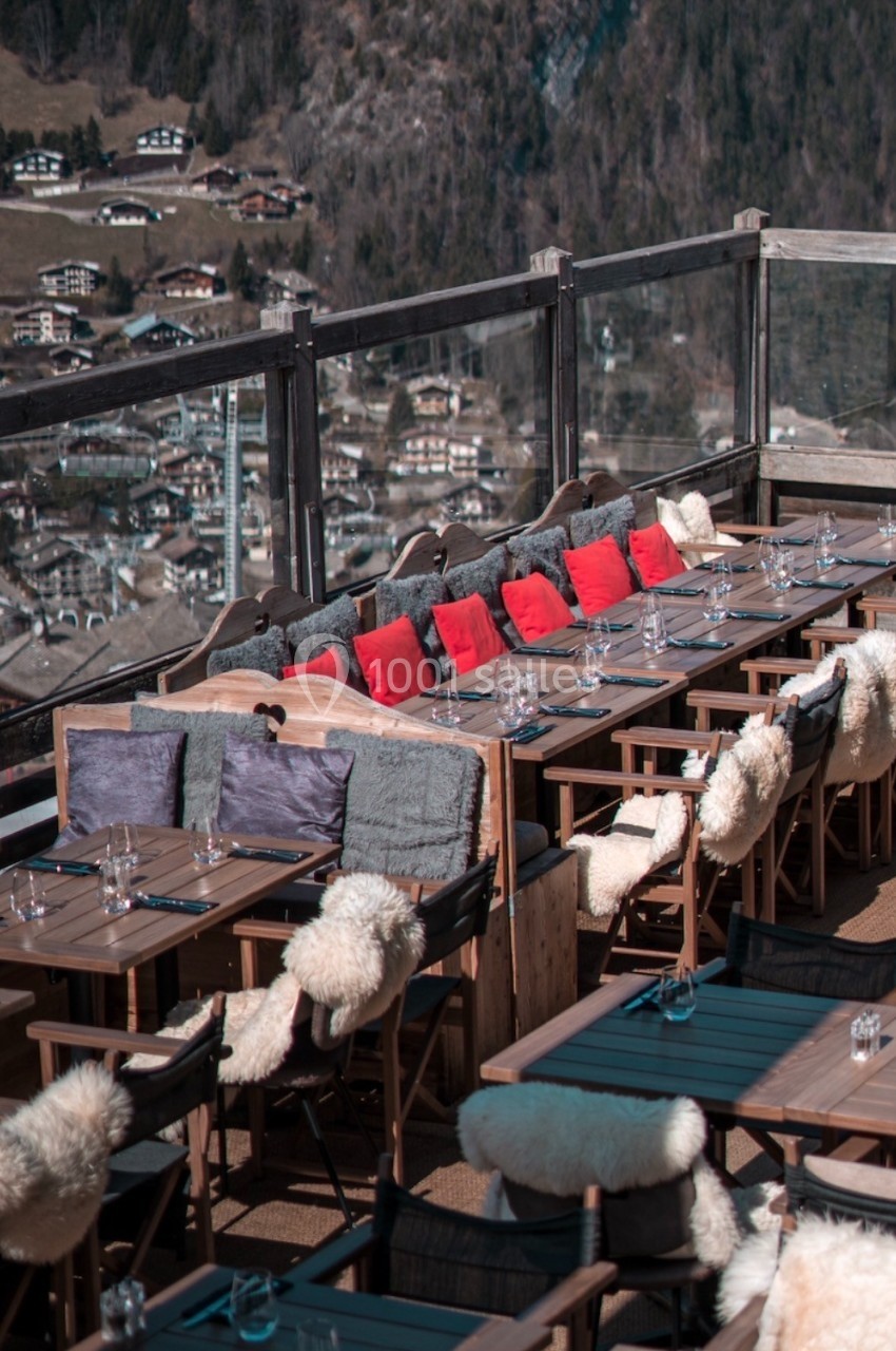 Terrasse en bois avec tables dressées, chaises garnies de coussins et vue sur un village de montagne en arrière-plan.