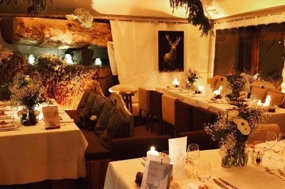 Une femme souriante se tient derrière une table en bois garnie de plats dans un chalet en bois éclairé chaleureusement.