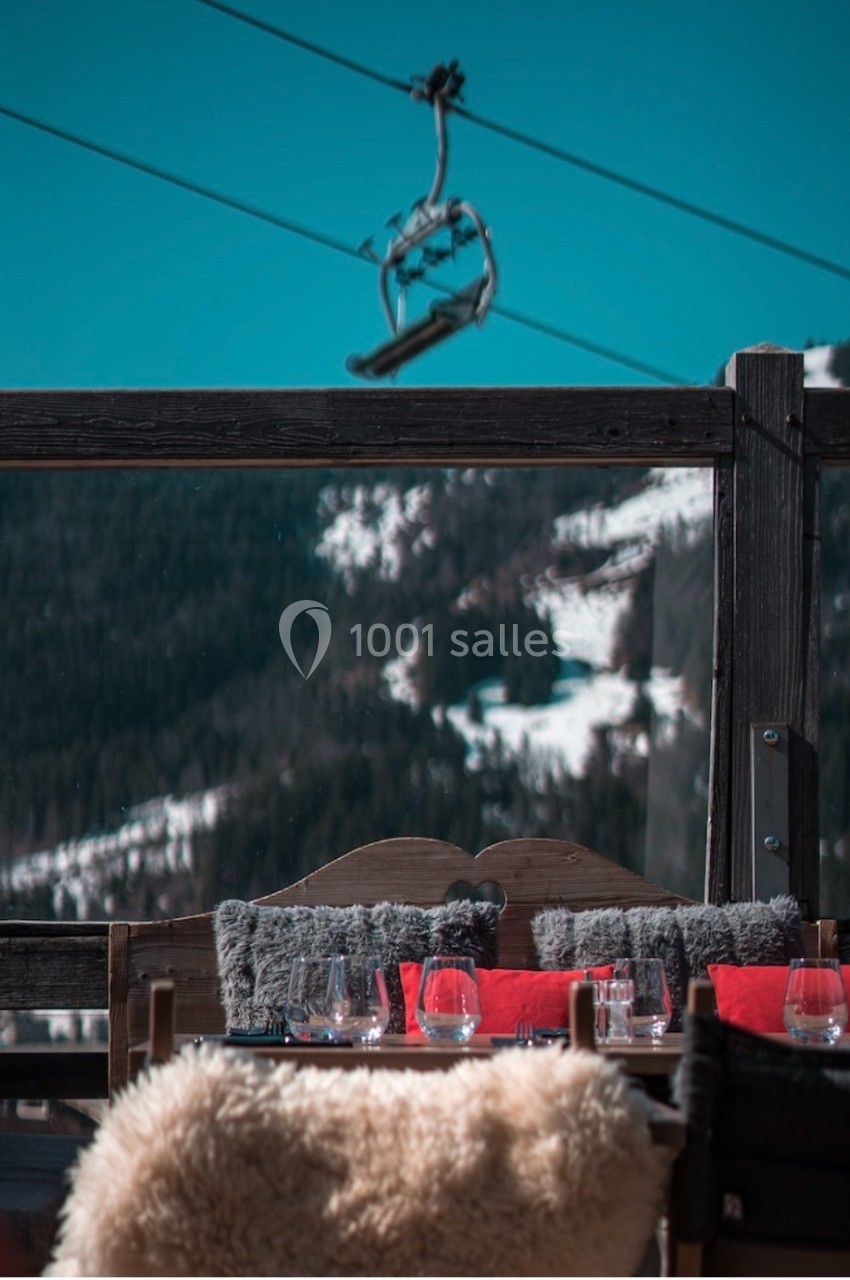 Terrasse en bois avec coussins et table dressée, vue sur des montagnes enneigées et un télésiège en arrière-plan.
