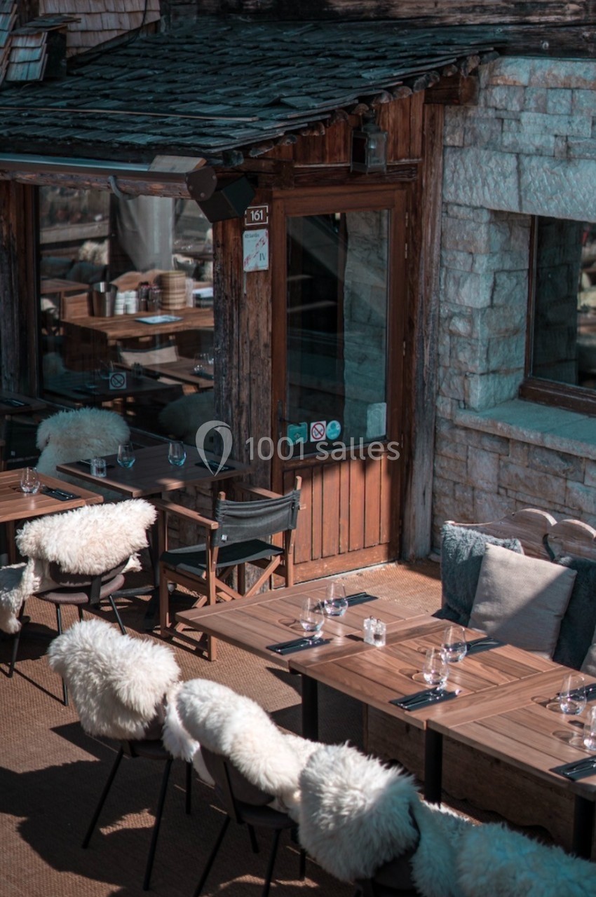 Terrasse en bois avec tables dressées, chaises garnies de fourrures blanches, devant une façade en pierre et bois.