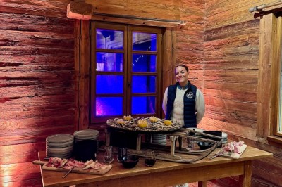 Une femme souriante se tient derrière une table en bois garnie de plats dans un chalet en bois éclairé chaleureusement.