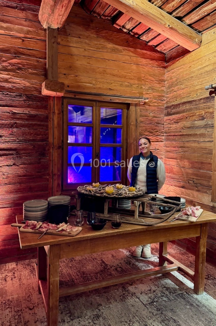 Une femme souriante se tient derrière une table en bois garnie de plats dans un chalet en bois éclairé chaleureusement.