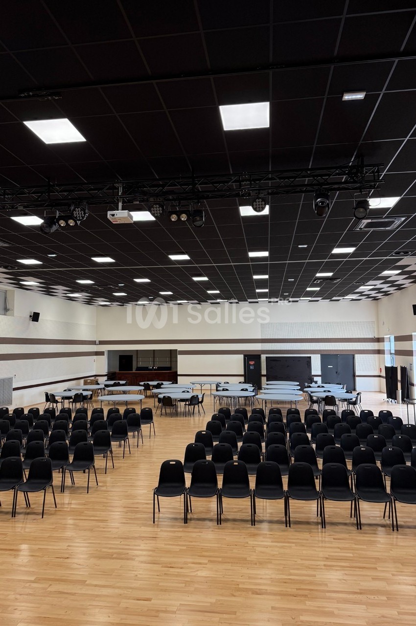 Salle polyvalente avec parquet, rangées de chaises noires, tables installées et plafond équipé de luminaires modernes.