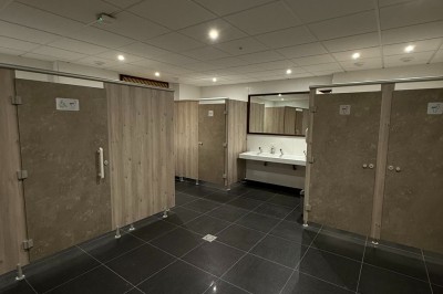 Miniature Location salle Erquy (Côtes-d'Armor) - Orion Evénement #8 Vue d'un espace de toilettes publiques avec cabines fermées, lavabos et miroir, sol en carrelage noir.