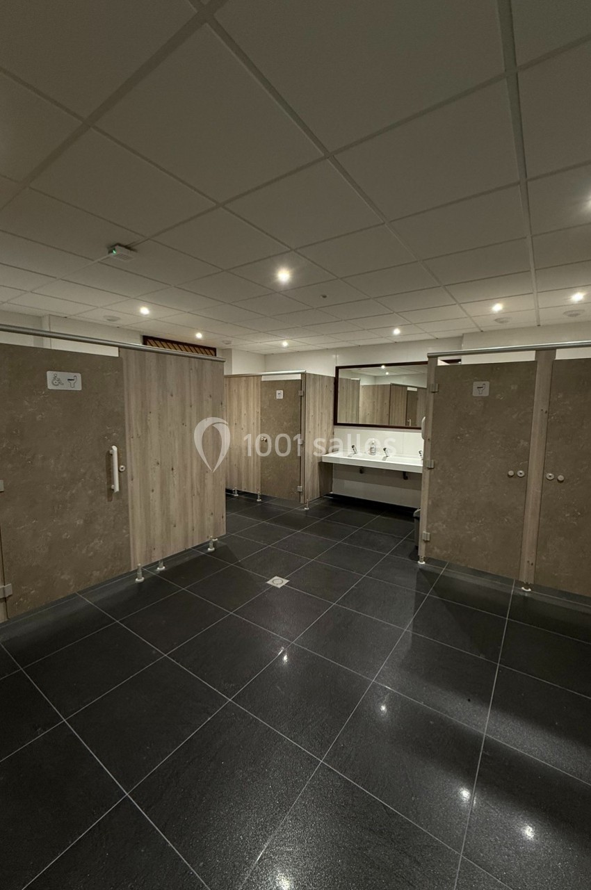 Vue d'un espace de toilettes publiques avec cabines fermées, lavabos et miroir, sol en carrelage noir.