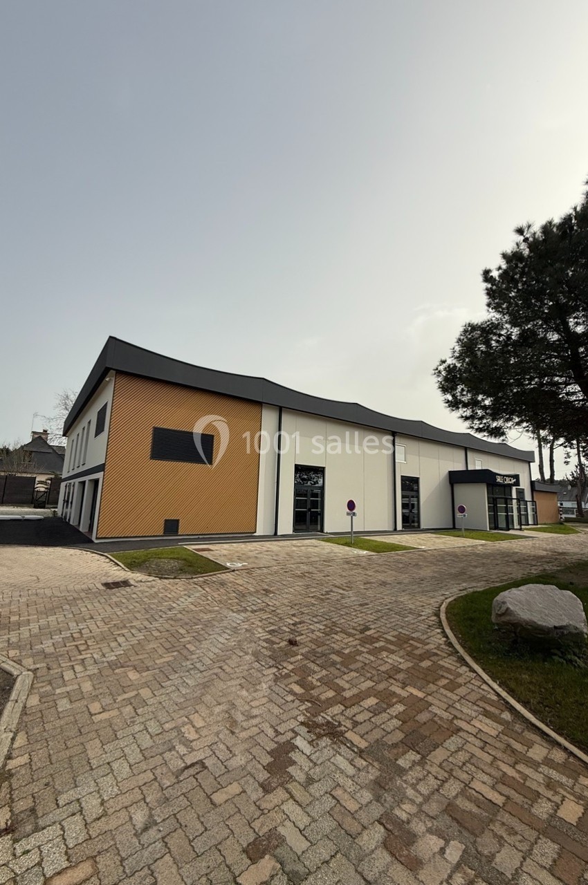 Bâtiment moderne avec façade beige et blanche, entouré d'un espace pavé et de pelouses avec quelques arbres.