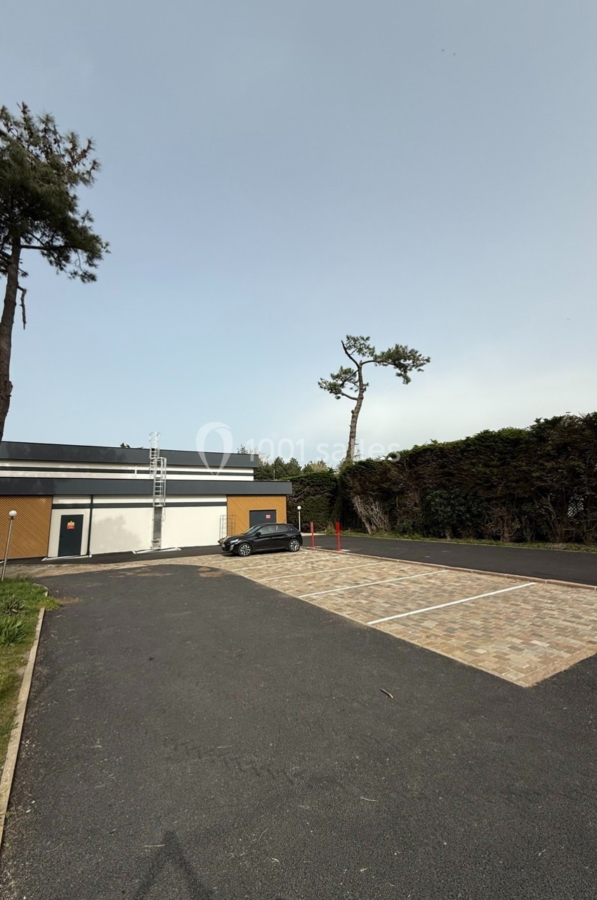 Parking vide avec une voiture noire stationnée près d'un bâtiment moderne, entouré de haies et d'arbres.