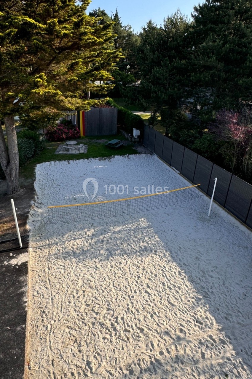 Terrain de sable avec un filet de volley, entouré de clôtures et d'arbres dans un espace extérieur.