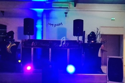 Salle décorée pour une fête avec des lumières colorées, une scène équipée de haut-parleurs et une bannière ’Happy Birthday’.