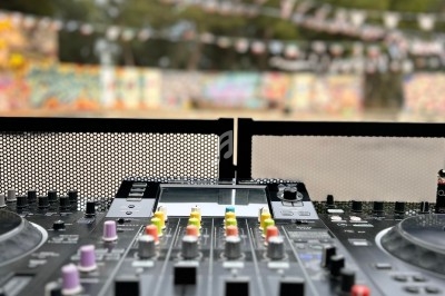 Table de mixage DJ avec curseurs et boutons colorés, en extérieur devant un décor flou de graffitis et arbres.