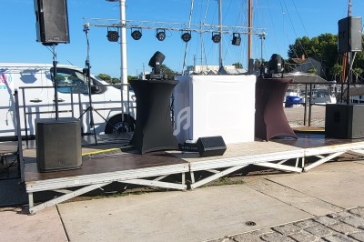 Scène extérieure avec équipements audio et éclairage, installée sur un quai près de voiliers sous un ciel bleu.