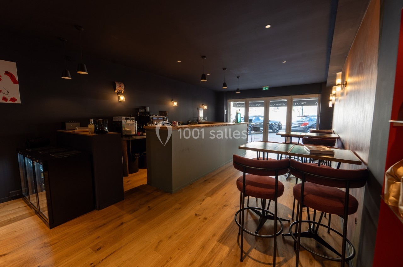Intérieur d'un café moderne avec comptoir en bois, tables hautes et chaises, éclairage tamisé et vue sur l'extérieur.