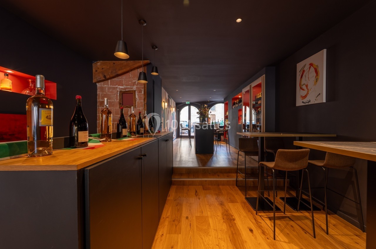 Intérieur d'un bar moderne avec comptoir en bois, bouteilles exposées et tables hautes dans une ambiance chaleureuse.