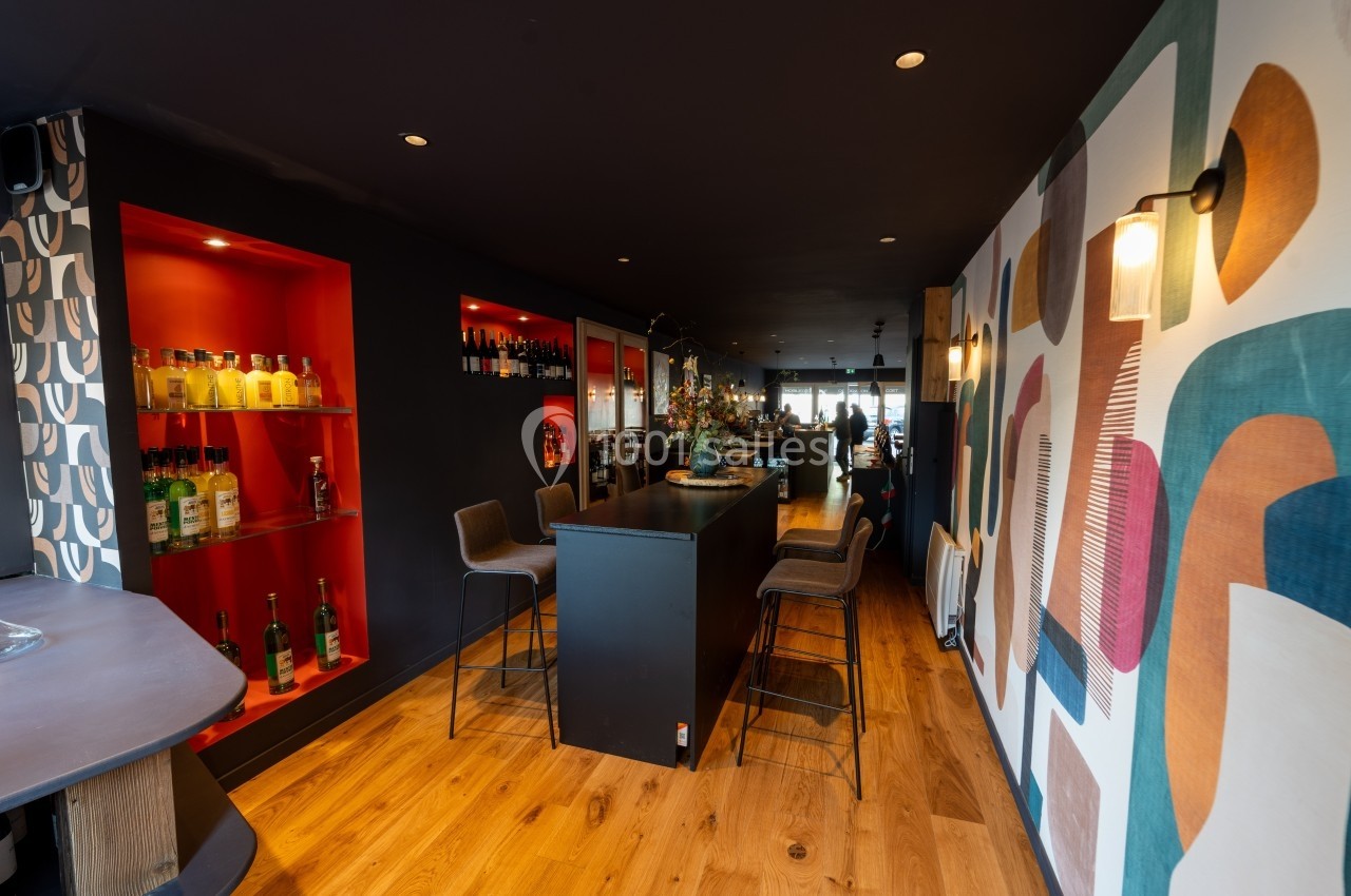 Intérieur d'un bar moderne avec comptoir central, étagères éclairées contenant des bouteilles et décoration murale colorée.