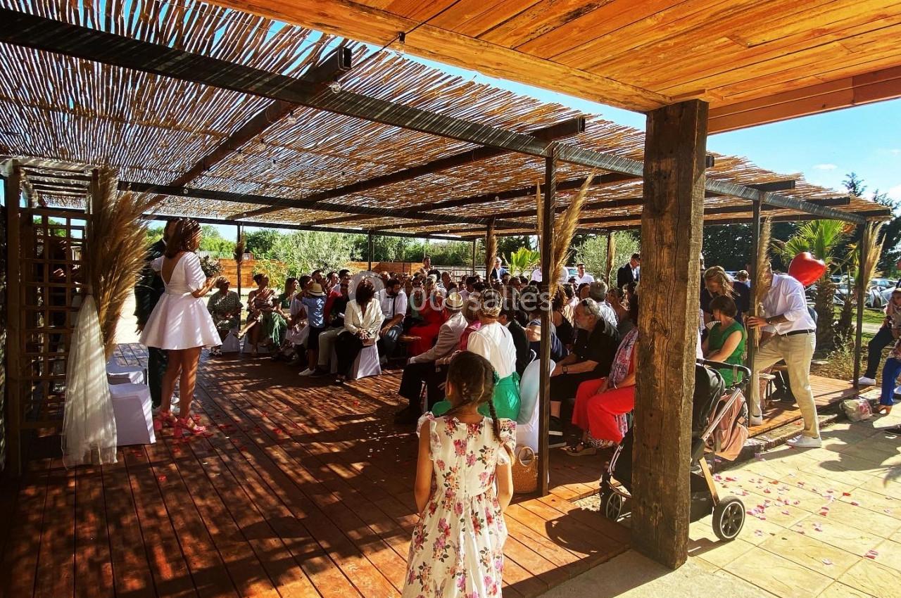 Une femme en robe blanche s'adresse à un public assis sous une pergola en bois lors d'un événement en extérieur.