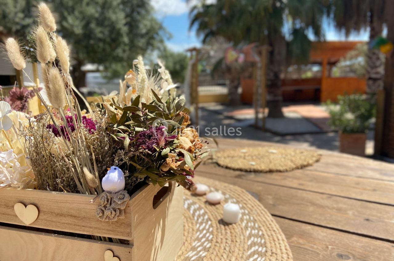 Centre de table en bois avec des fleurs séchées, posé sur une table en bois dans un espace extérieur ensoleillé.