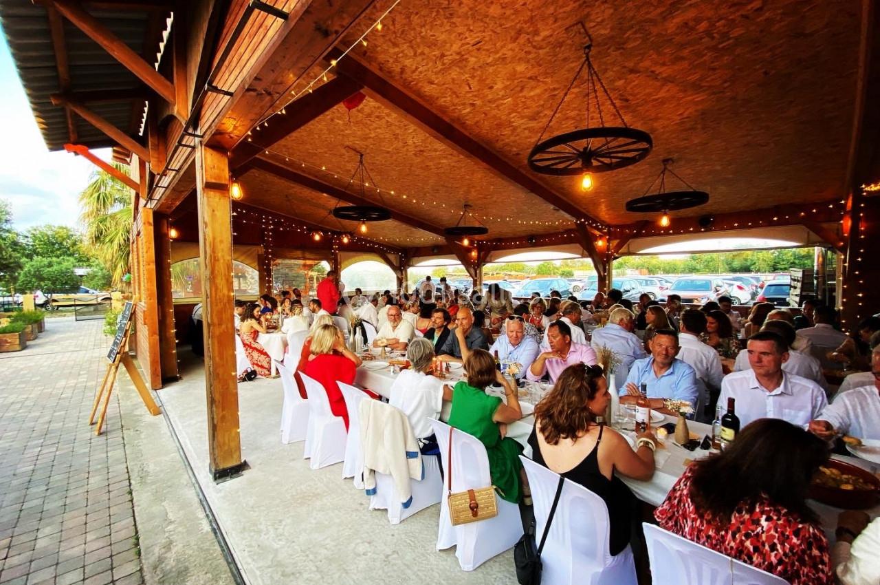 Des convives assis à de longues tables sous un auvent en bois, décoré de lumières suspendues, lors d'un événement festif.