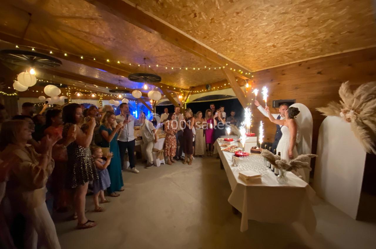 Une mariée allume des cierges magiques sur un gâteau devant des invités applaudissant dans une salle décorée.