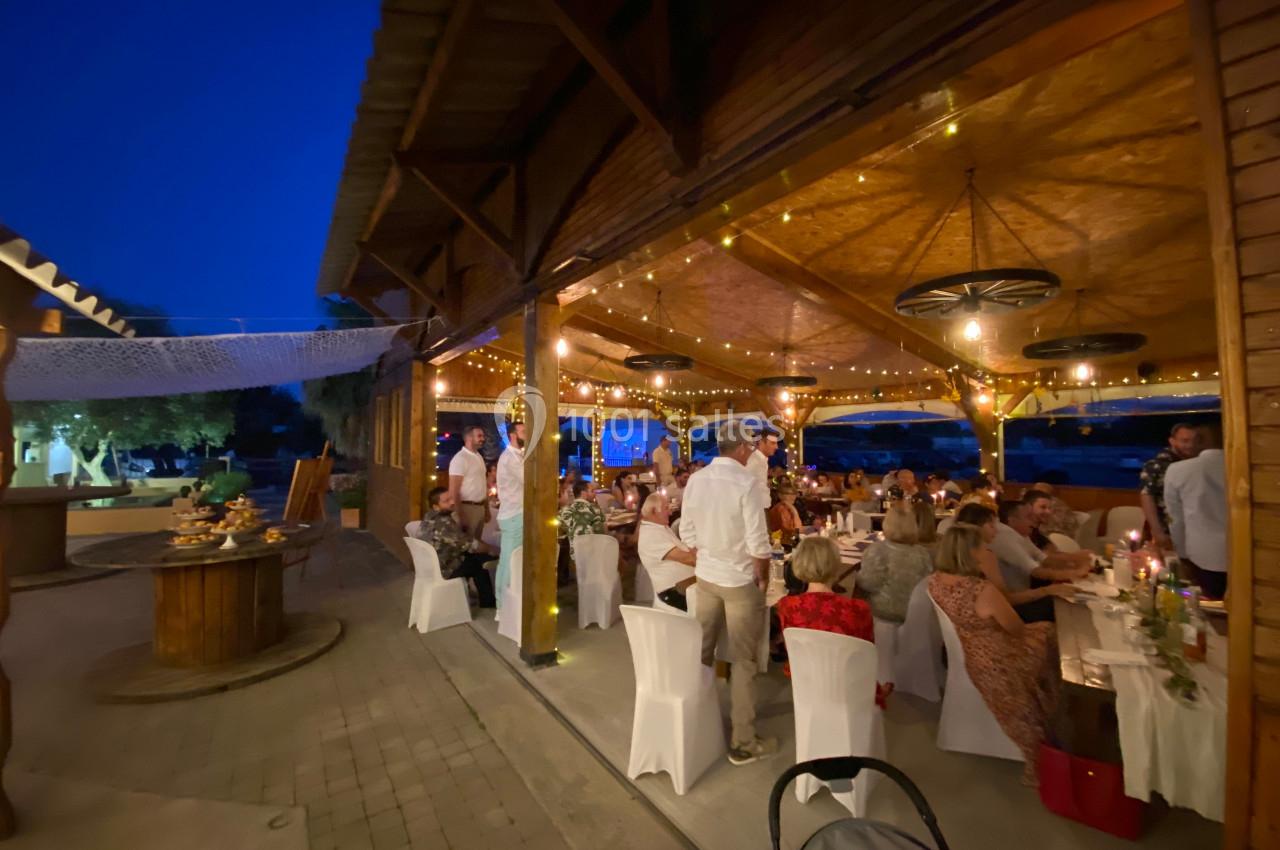 Salle de réception en soirée avec des invités assis à des tables décorées, éclairée par des guirlandes lumineuses.