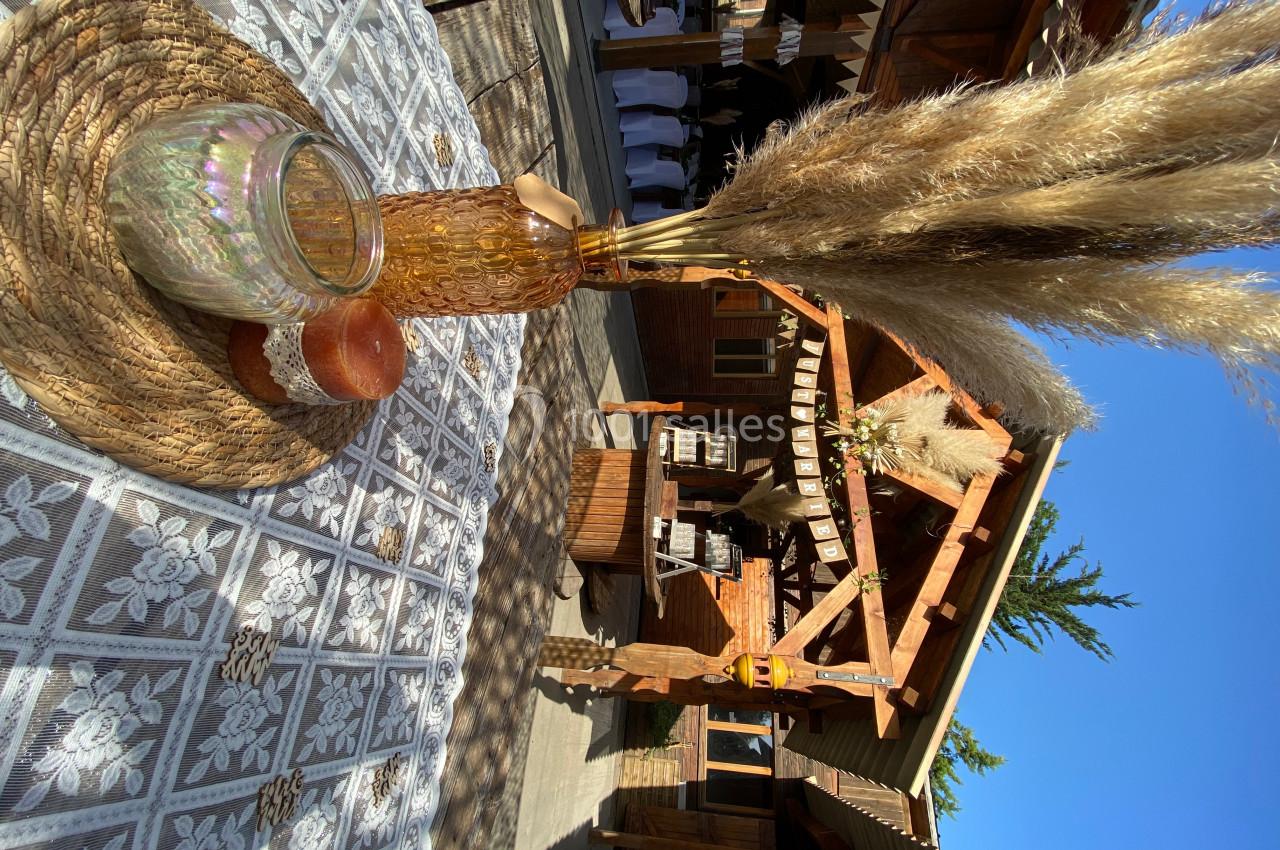 Centre de table avec vase, pampas et bougies sur nappe en dentelle, devant un bâtiment en bois sous un ciel bleu.