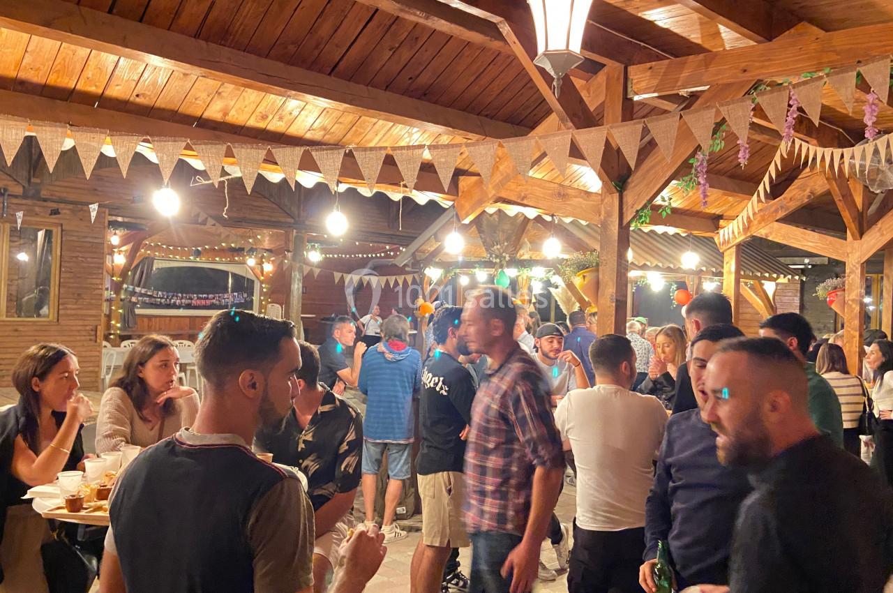 Groupe de personnes discutant et mangeant dans un espace en bois décoré de guirlandes et éclairé par des lampes suspendues.