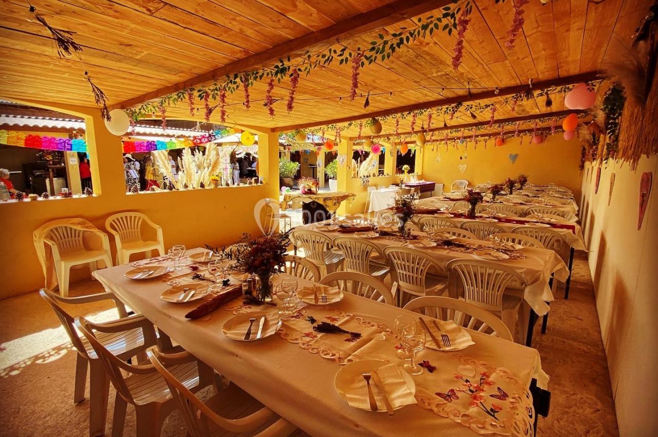 Salle décorée avec des tables alignées, nappes blanches, chaises en plastique et ornements suspendus sous un toit en bois.