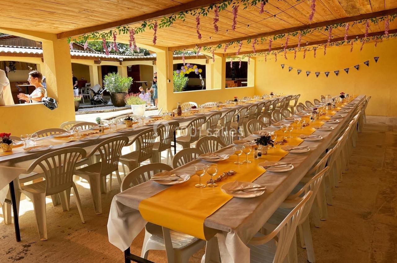 Tables longues décorées avec nappes jaunes et blanches, disposées sous un auvent en bois pour un repas en extérieur.