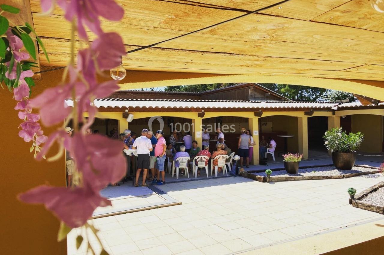 Groupe de personnes assises et debout sous un auvent dans une cour ensoleillée, avec des fleurs en premier plan.