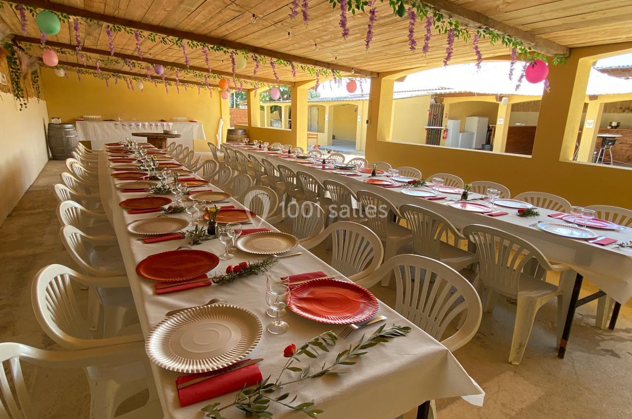 Salle décorée avec des tables alignées, nappées de blanc, ornées d'assiettes colorées et de fleurs, sous un toit en bois.