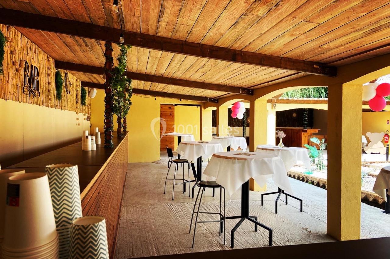 Terrasse couverte avec tables hautes dressées, bar en bois à gauche et décorations simples en arrière-plan.