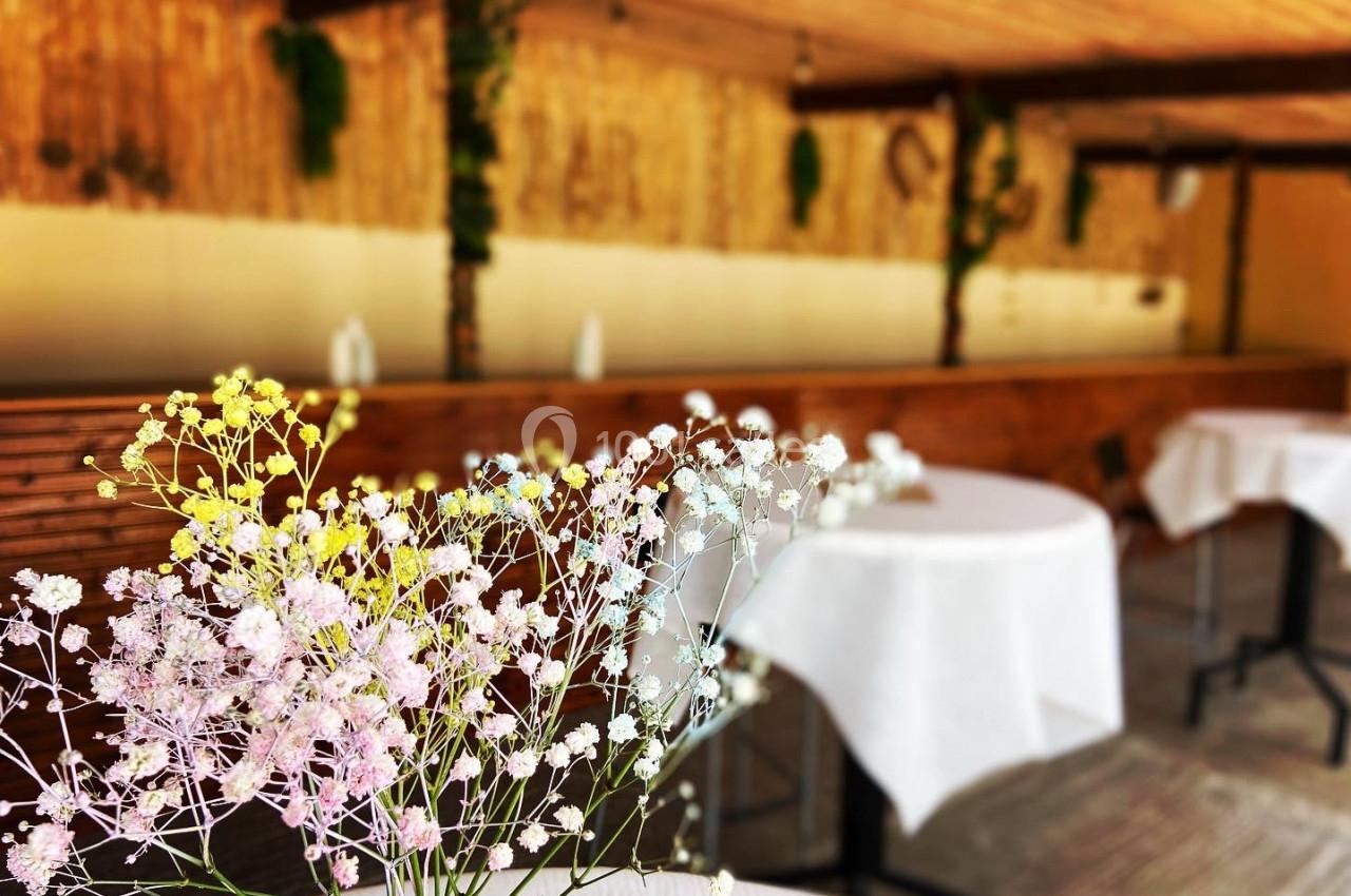 Fleurs délicates au premier plan avec une salle en bois, des tables rondes et une ambiance chaleureuse en arrière-plan.