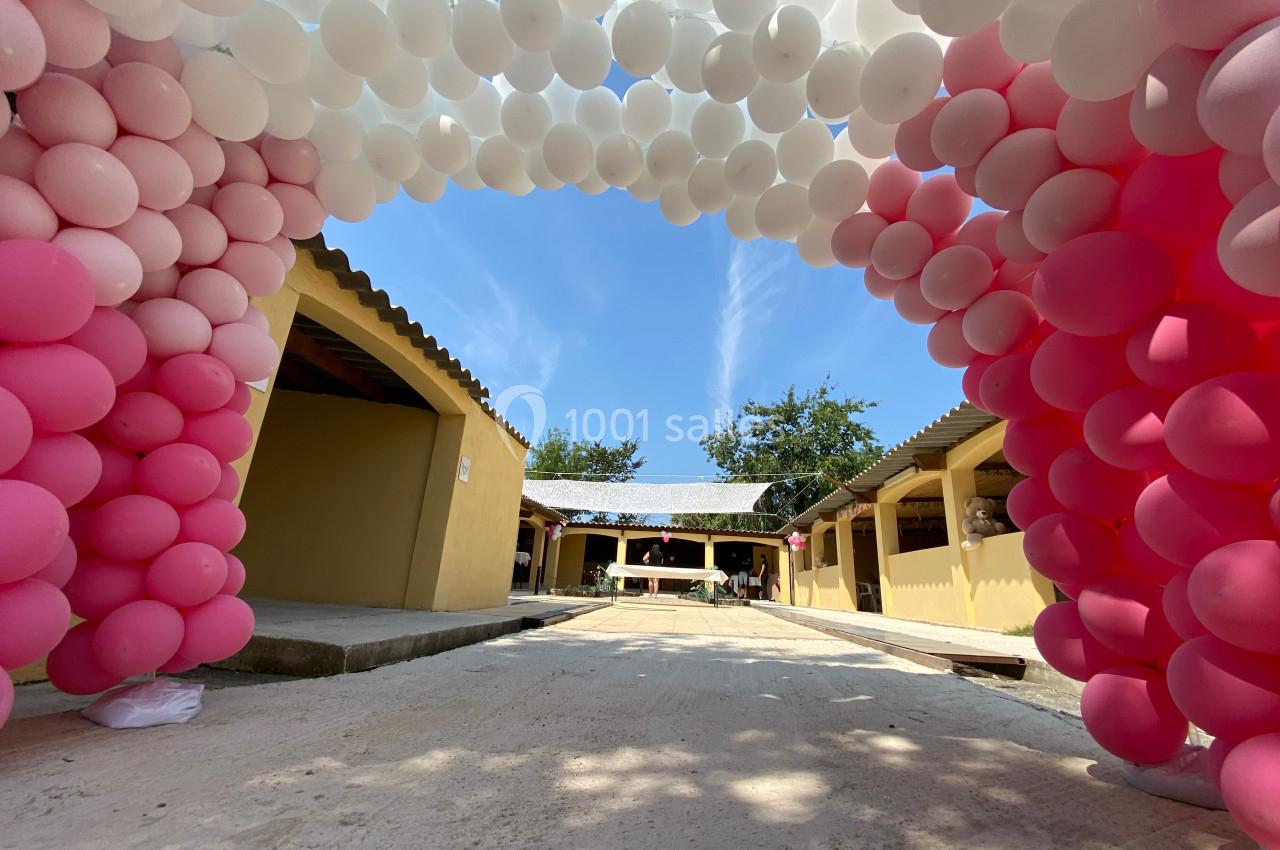 Arche de ballons roses et blancs décorant une allée extérieure bordée de bâtiments jaunes sous un ciel dégagé.
