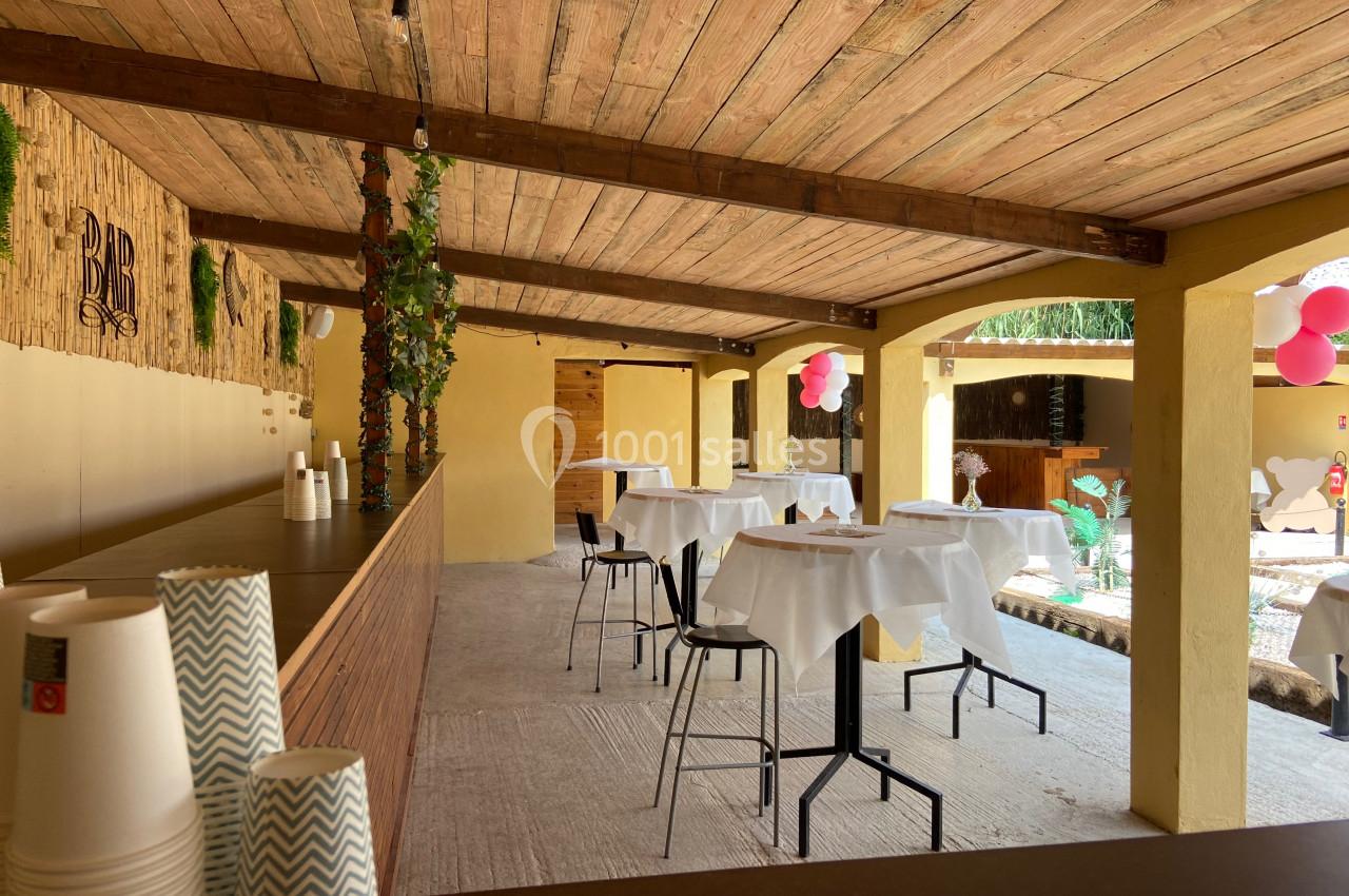 Salle extérieure couverte avec tables dressées, bar en bois, décorations végétales et ballons roses.