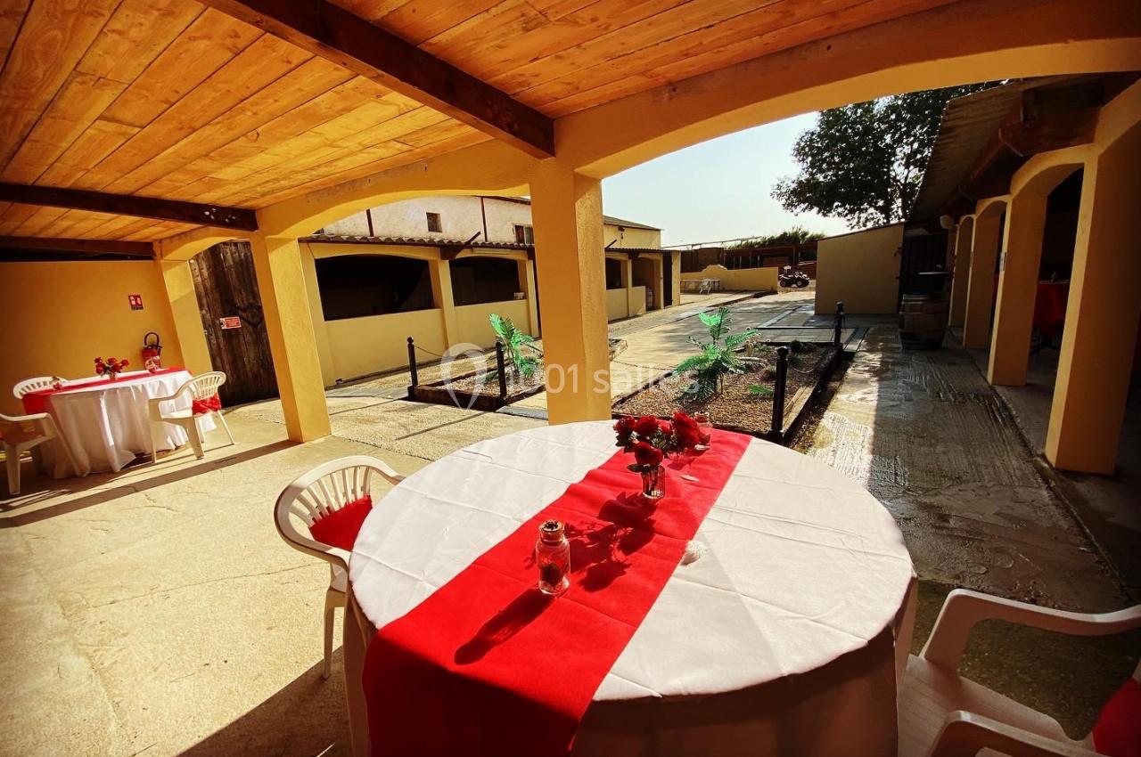 Table ronde décorée d'une nappe blanche et rouge sous un auvent en bois, avec vue sur une cour ensoleillée.