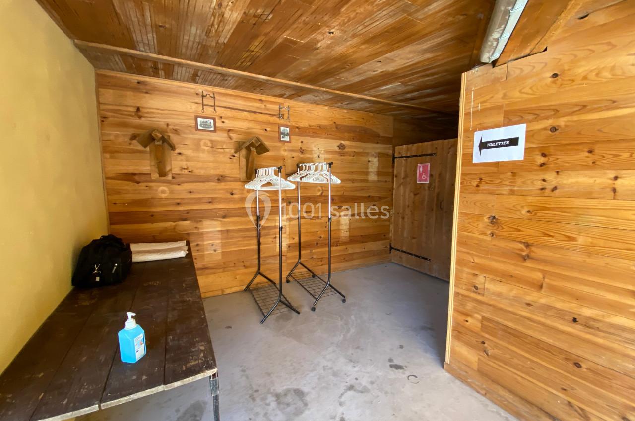 Vestiaire en bois avec porte-manteaux, table, gel désinfectant et signalisation des toilettes sur la porte.