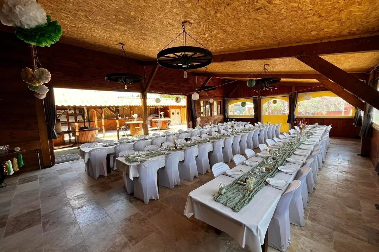 Salle de réception en bois avec deux longues tables dressées, décorations suspendues et lumière naturelle.