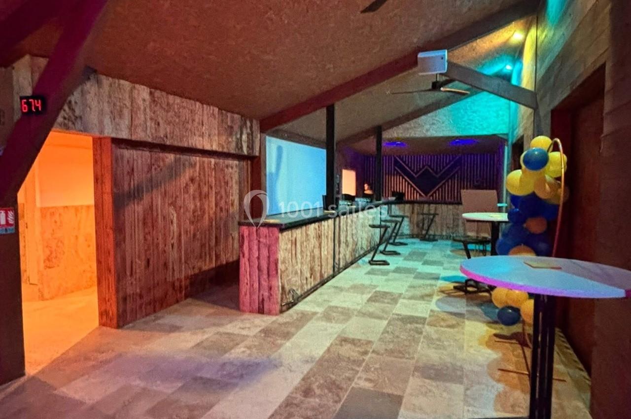 Salle avec un comptoir en bois, éclairage coloré, tables hautes et décorations en ballons jaunes et bleus.