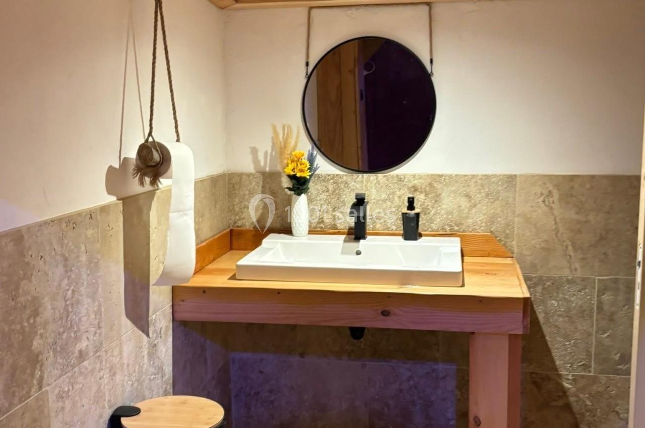 Lavabo en bois avec miroir rond, vase décoratif et poubelle noire dans une salle de bain au plafond en bois.
