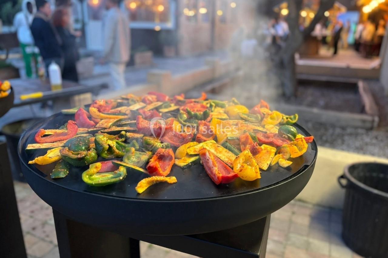 Légumes colorés grillant sur une plancha en extérieur, avec des convives flous en arrière-plan.