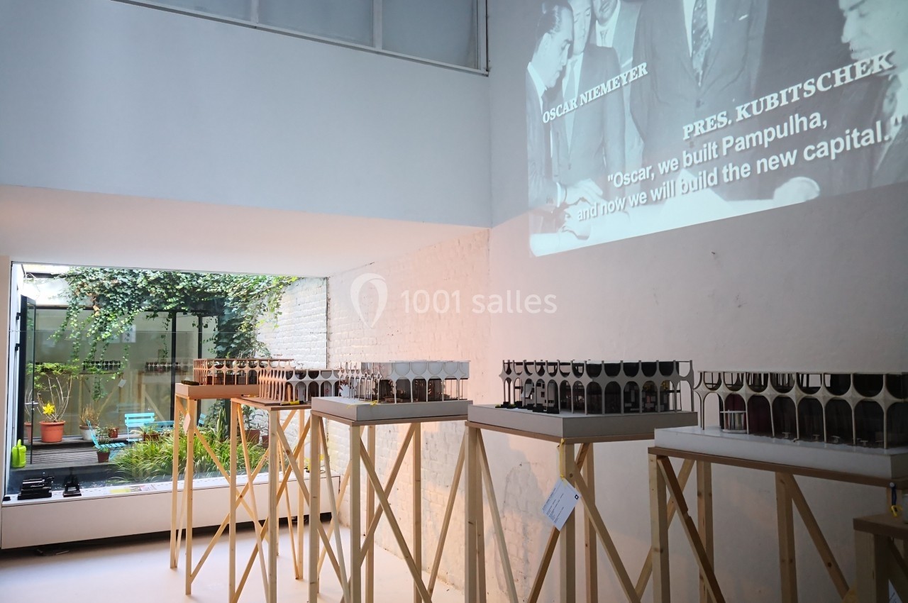 Exposition de maquettes architecturales sur des supports en bois dans une salle lumineuse avec une projection murale.